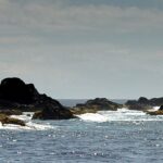 Panorama der Kette schwarzer Klippen der Formigas knapp über dem Wasser, mit weißer Gischt gesäumt, auf glitzernd blauem Meer, rechts der weiße Leuchtturm von Formigão unter dunstigem Himmel