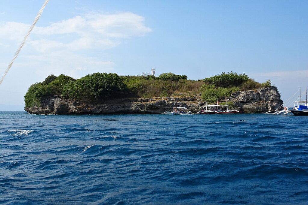 Die Insel Pescador vom Meer aus, mit grünem Buschwerk bewachsen, oben der weiße Leuchtturm und am Fuß der Klippe drei festgemachte Tauchbancas
