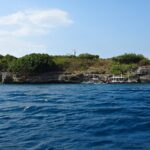 Die Insel Pescador vom Meer aus, mit grünem Buschwerk bewachsen, oben der weiße Leuchtturm und am Fuß der Klippe drei festgemachte Tauchbancas
