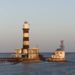 Der Leuchtturm von Daedalus, ein schwarz-weiß gestreifter Turm auf dem Riff, mit seinen Gebäuden und dem Metallsteg, bei Sonnenuntergang