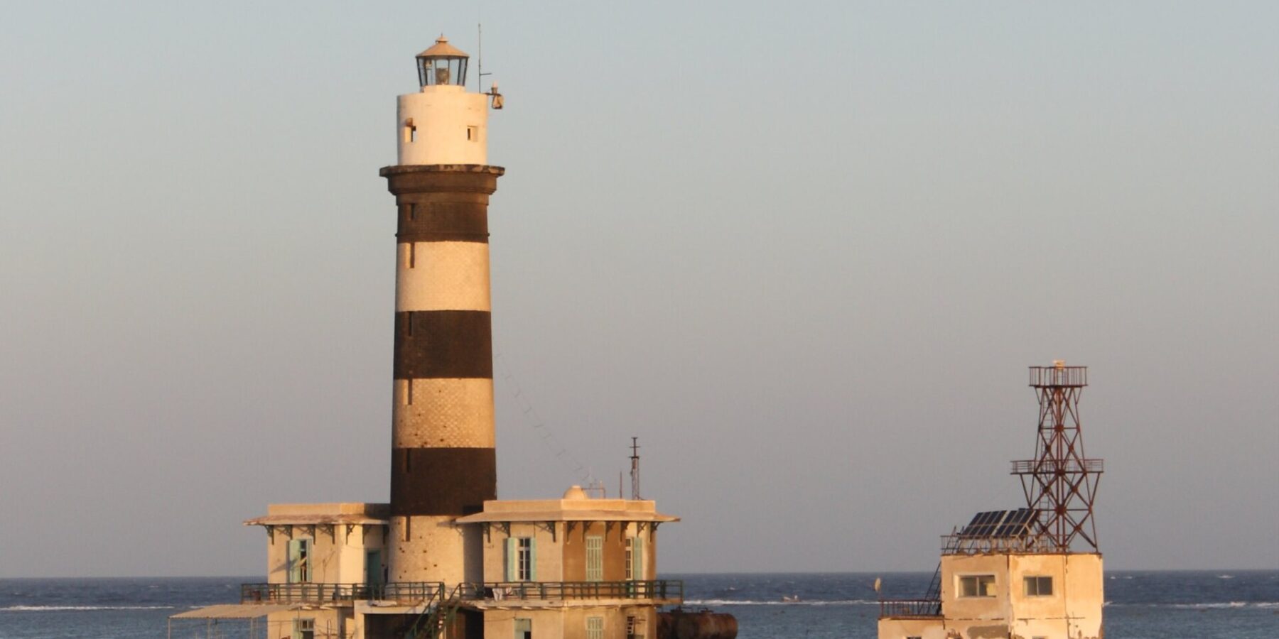 Der Leuchtturm von Daedalus, ein schwarz-weiß gestreifter Turm auf dem Riff, mit seinen Gebäuden und dem Metallsteg, bei Sonnenuntergang
