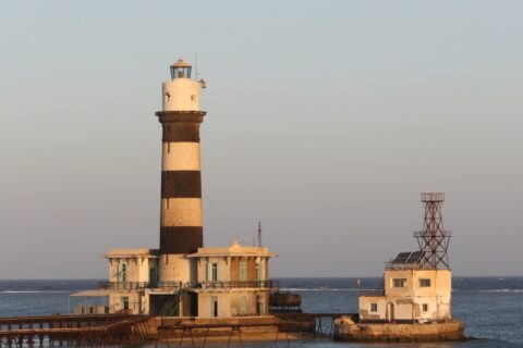 Der Leuchtturm von Daedalus, ein schwarz-weiß gestreifter Turm auf dem Riff, mit seinen Gebäuden und dem Metallsteg, bei Sonnenuntergang