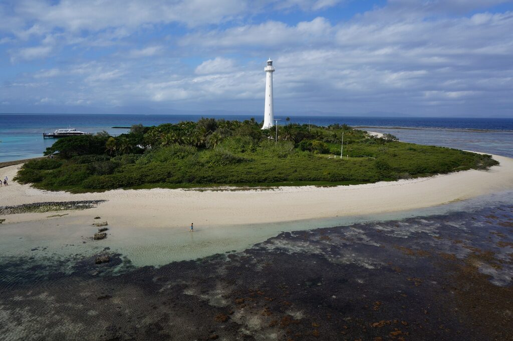 Der große weiße Leuchtturm der Insel Amédée ragt über die Vegetation, die Insel ist von weißem Sand umgeben, im Vordergrund das trockengefallene Riffdach und links ein Boot am Anleger