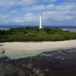 Der große weiße Leuchtturm der Insel Amédée ragt über die Vegetation, die Insel ist von weißem Sand umgeben, im Vordergrund das trockengefallene Riffdach und links ein Boot am Anleger