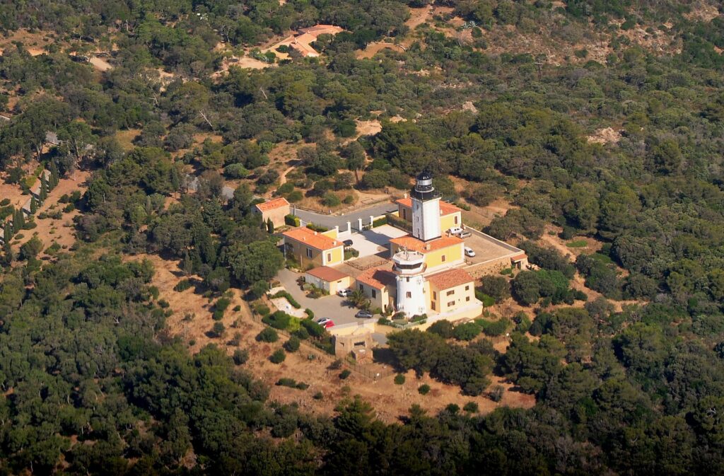Luftaufnahme des Leuchtturms am Cap Camarat, ein weißer Turm mit schwarzer Laterne, umgeben von gelben Gebäuden mit orangefarbenen Ziegeldächern, inmitten eines dichten mediterranen Waldes.