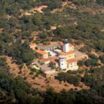 Luftaufnahme des Leuchtturms am Cap Camarat, ein weißer Turm mit schwarzer Laterne, umgeben von gelben Gebäuden mit orangefarbenen Ziegeldächern, inmitten eines dichten mediterranen Waldes.