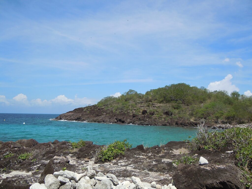 Eine türkise Bucht, geschützt von einer dunklen, buschbewachsenen Felsspitze, von den Felsen der Insel aus gesehen, unter blauem Himmel