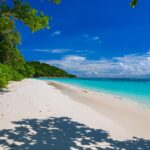 Der weiße Sandstrand von Koh Tachai, von Bäumen gesäumt, unter blauem Himmel, rechts türkises Wasser und draußen eine Reihe Bojen