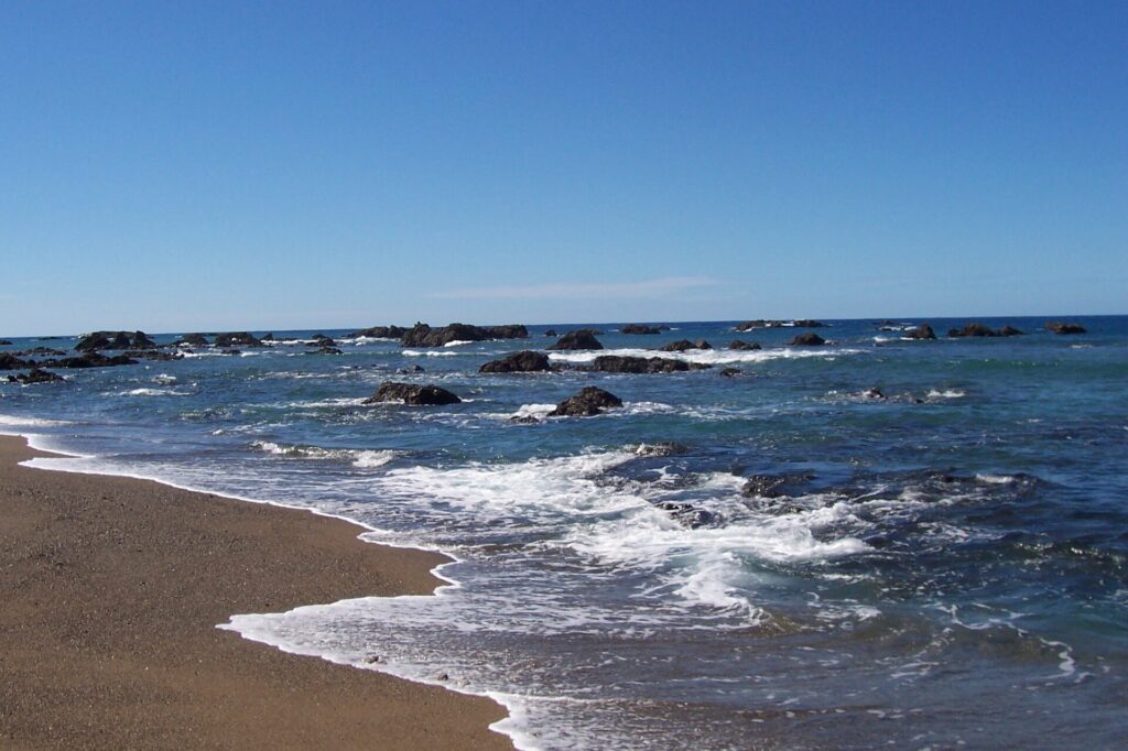 Ein goldbrauner Sandstrand, an dem schaumgesäumte Wellen auslaufen, vor einem blauen Meer mit schwarzen Felsen bis zum Horizont, unter wolkenlosem Himmel.