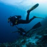 Zwei Taucher über einem Riffhang, einer an der Leine eines Riffhakens, der andere in Shorts, in blauem Wasser voller kleiner Fische