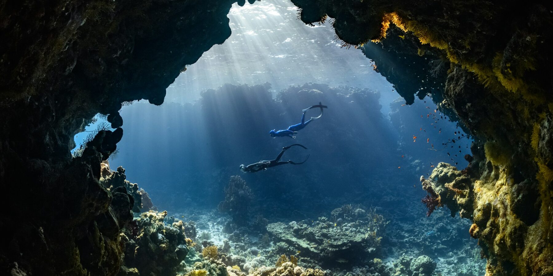 Zwei Taucher schwimmen durch eine Unterwasserhöhle, die von Sonnenstrahlen erhellt wird, über einem Riff