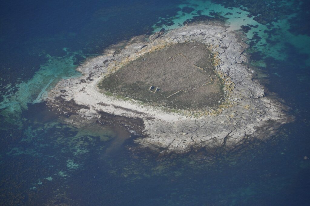 Luftbild einer kleinen Insel aus grauen Granitplatten, in der Mitte mit trockenem Gras bewachsen, in dem sich verfallene Mäuerchen abzeichnen, gesäumt von hellem Strand und umgeben von türkisen Untiefen und tiefblauem Wasser.