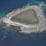 Luftbild einer kleinen Insel aus grauen Granitplatten, in der Mitte mit trockenem Gras bewachsen, in dem sich verfallene Mäuerchen abzeichnen, gesäumt von hellem Strand und umgeben von türkisen Untiefen und tiefblauem Wasser.