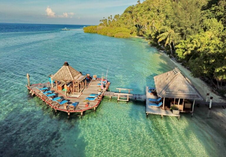 Eine Plattform auf Pfählen mit blauen Liegen, über einen Steg mit einem Pavillon am Strand verbunden