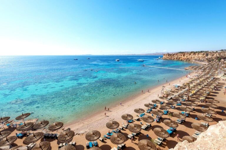 Ein langer Sandstrand am Fuß einer Klippe, mit Strohschirmen und einem Steg über dem türkisfarbenen Meer