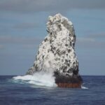 Die Nordwestspitze der Roca Partida, weiß vom Guano, vom Boot aus gesehen, eine Welle bricht an ihrem Fuß, darüber fliegende Vögel