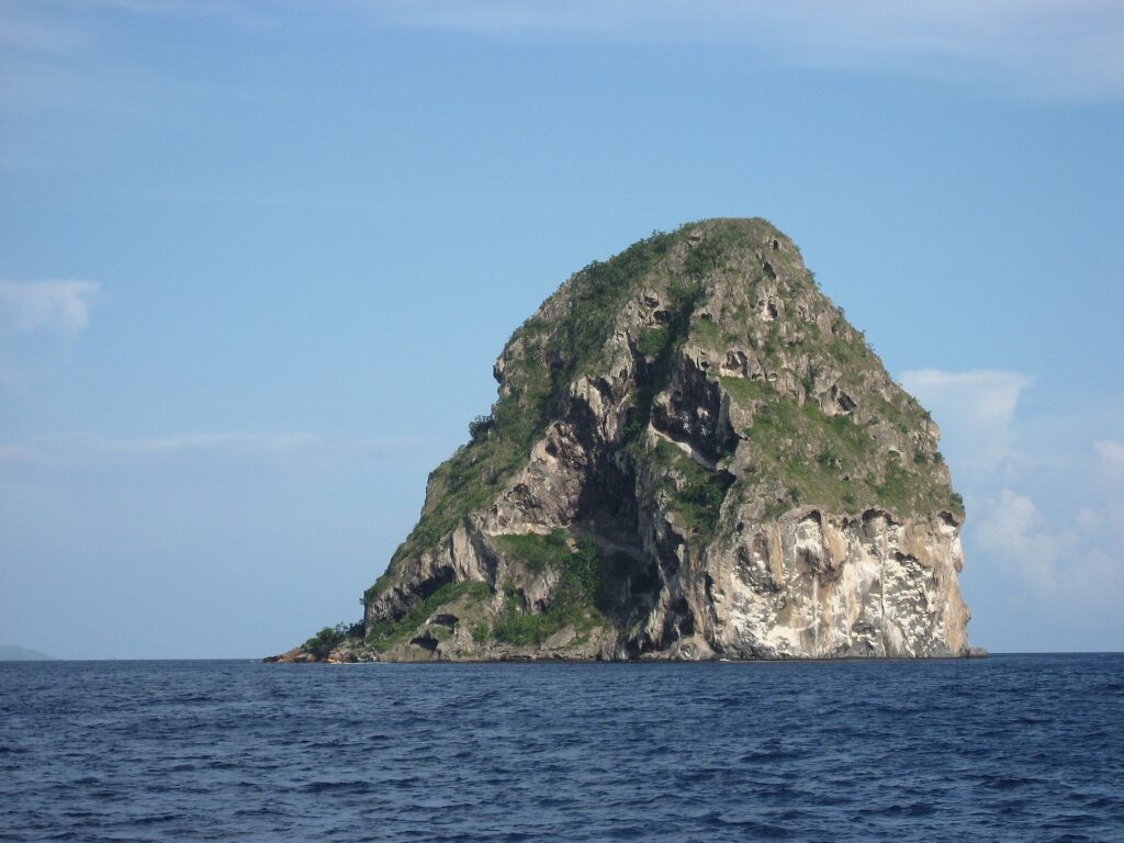 Der Diamantfelsen aus der Nähe vom Meer aus, eine graue Felsmasse mit Höhlen, weiß gestreift und von Vegetation gekrönt, über dunkelblauem Meer