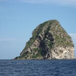 Der Diamantfelsen aus der Nähe vom Meer aus, eine graue Felsmasse mit Höhlen, weiß gestreift und von Vegetation gekrönt, über dunkelblauem Meer