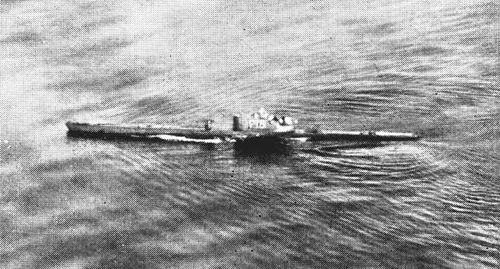 Schwarz-Weiß-Archivfoto aus einem Flugzeug: Ein U-Boot fährt an der Oberfläche über ein gekräuseltes Meer, mit Kielwasser am Rumpf und dem Turm in der Mitte.