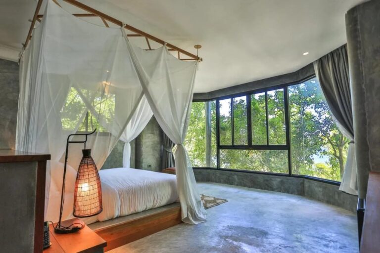 Ein Zimmer mit Betonwänden, einem Bett unter Moskitonetz und einer Glasfront zu den Bäumen