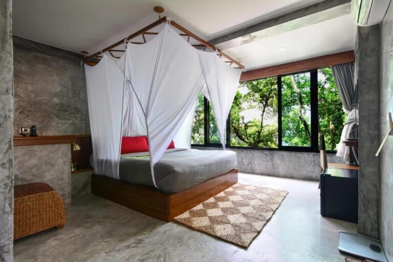 Ein Bett unter Moskitonetz mit roten Kissen, vor einem großen Fenster zum Wald