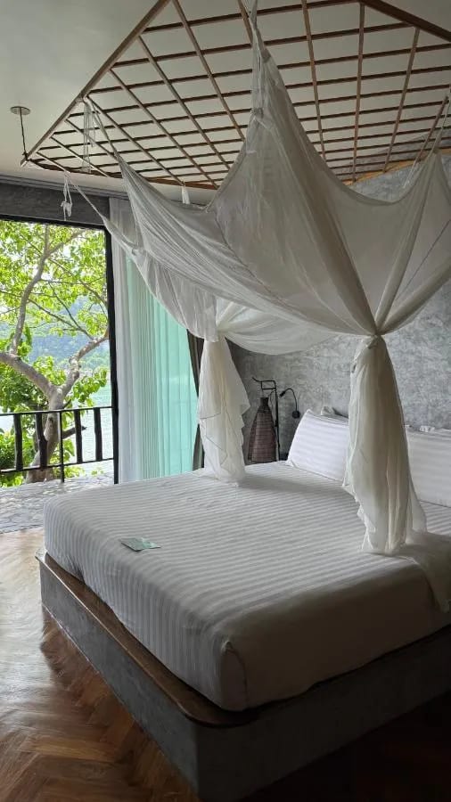 Ein Bett unter Moskitonetz vor einer Glastür, mit Balkon und Blick auf die Bäume und das Meer