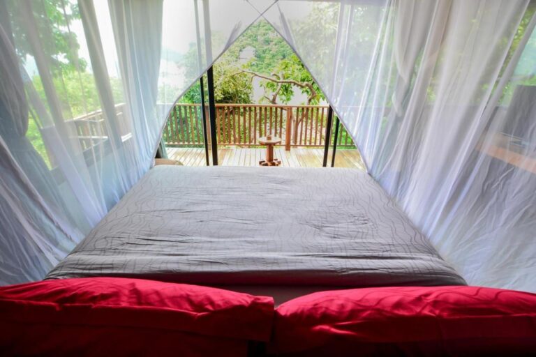 Blick vom Bett durch das Moskitonetz auf eine Holzterrasse und die Bäume