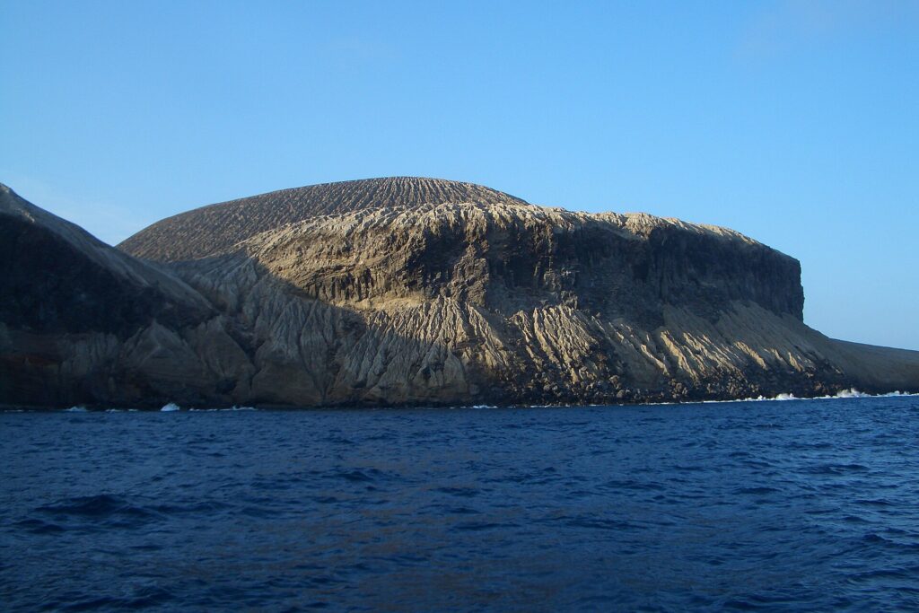 Die Küste von San Benedicto vom Meer aus: der von Rinnen durchzogene Aschekegel des Vulkans Bárcena über dunklen Klippen und tiefblauem Meer