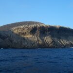 Die Küste von San Benedicto vom Meer aus: der von Rinnen durchzogene Aschekegel des Vulkans Bárcena über dunklen Klippen und tiefblauem Meer