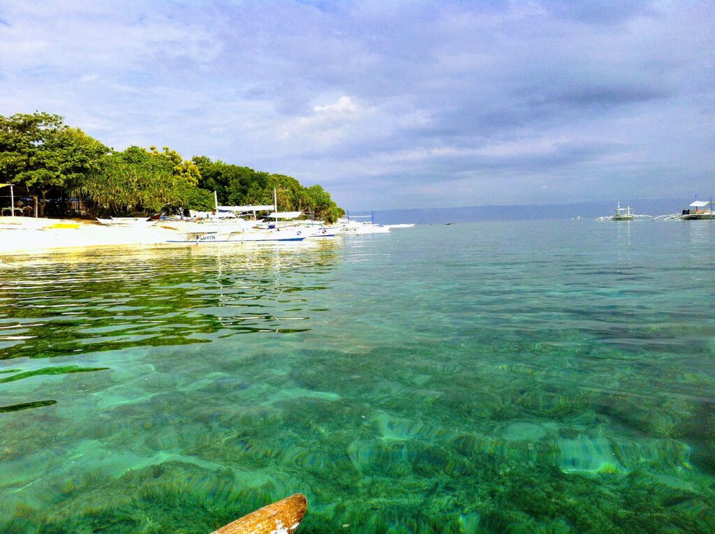 Klares türkisfarbenes Wasser über flachem Grund vor einem weißen Sandstrand mit Bäumen, an dem weiße Bangkas aufgereiht liegen, weitere Boote draußen unter bewölktem Himmel.