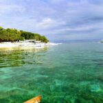 Klares türkisfarbenes Wasser über flachem Grund vor einem weißen Sandstrand mit Bäumen, an dem weiße Bangkas aufgereiht liegen, weitere Boote draußen unter bewölktem Himmel.