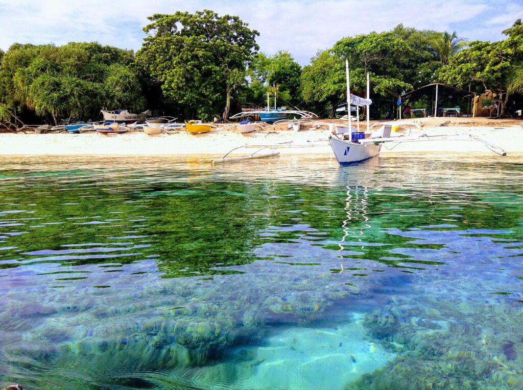 Knapp über der Wasserlinie: Korallenblöcke unter klarem türkisfarbenem Wasser, vor einem weißen Sandstrand mit einer ankernden weißen Bangka, weitere bunte Boote unter die Bäume gezogen.