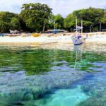Knapp über der Wasserlinie: Korallenblöcke unter klarem türkisfarbenem Wasser, vor einem weißen Sandstrand mit einer ankernden weißen Bangka, weitere bunte Boote unter die Bäume gezogen.