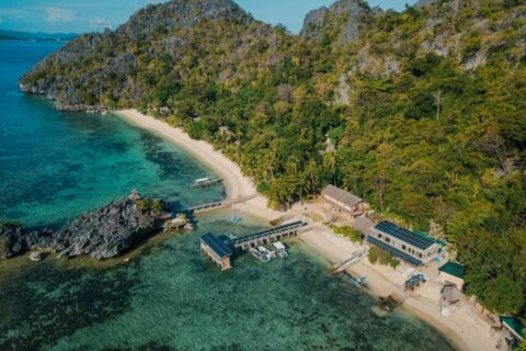 Luftaufnahme des Sangat Island Dive Resorts mit Strand, Steg und Booten am Fuß der Klippen