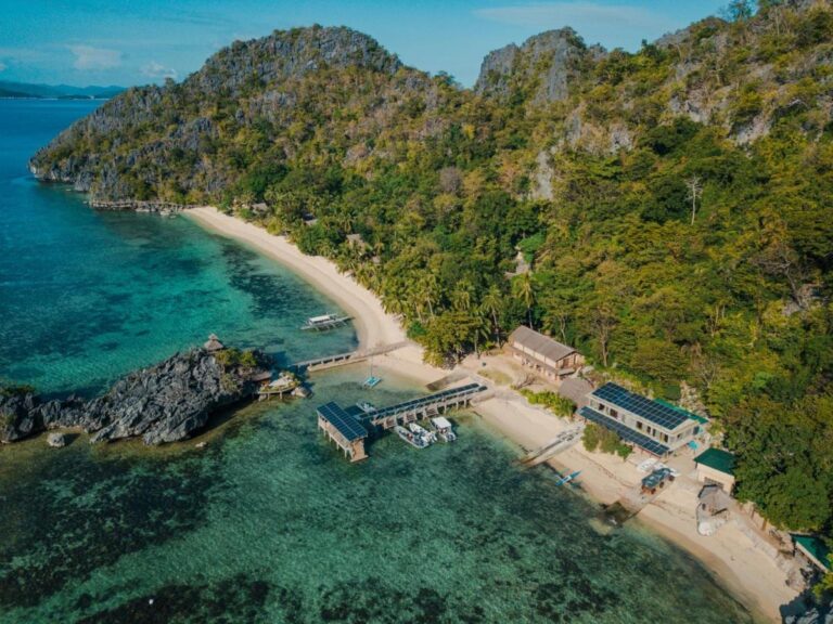 Luftaufnahme des Sangat Island Dive Resorts mit Strand, Steg und Booten am Fuß der Klippen