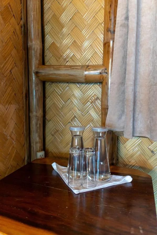 Zwei Wasserkaraffen und Gläser auf einem Holztisch vor einer Wand aus geflochtenem Bambus