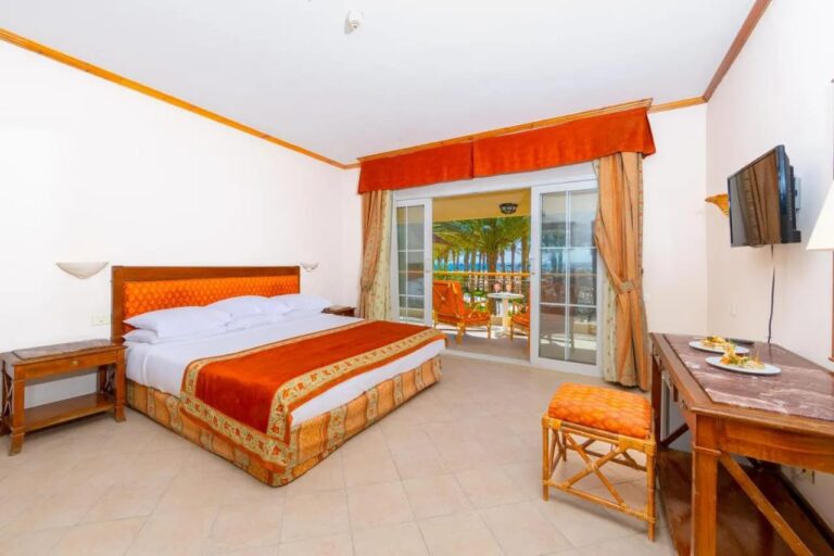 Ein Zimmer mit Doppelbett, offen zum Balkon, im Hintergrund Palmen und das Meer