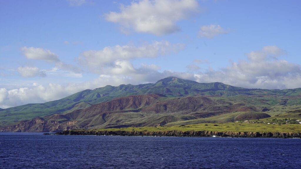 Die Küste der Insel Socorro vom Meer aus: schwarze Lavaklippen, grüne und rotbraune Hügel und der Gipfel des Vulkans in den Wolken