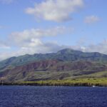 Die Küste der Insel Socorro vom Meer aus: schwarze Lavaklippen, grüne und rotbraune Hügel und der Gipfel des Vulkans in den Wolken