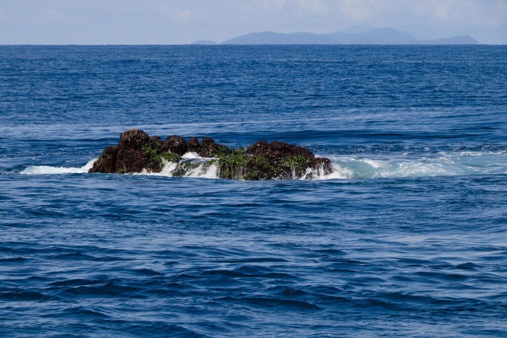 Der Gipfel des Richelieu Rock ragt bei Ebbe heraus, ein dunkler, mit grünen Algen bedeckter Felsen in der Brandung, die Surin-Inseln am Horizont