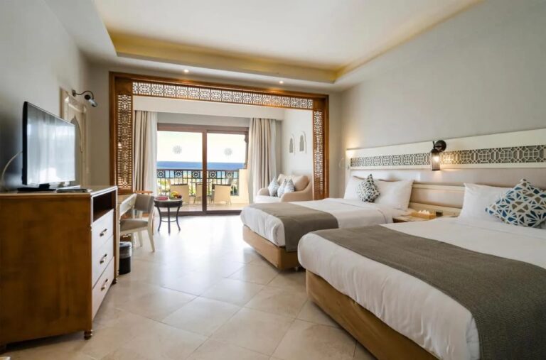 Ein Zimmer mit zwei Doppelbetten, einer Sitzecke unter einem Holzbogen und einem Balkon zum Meer