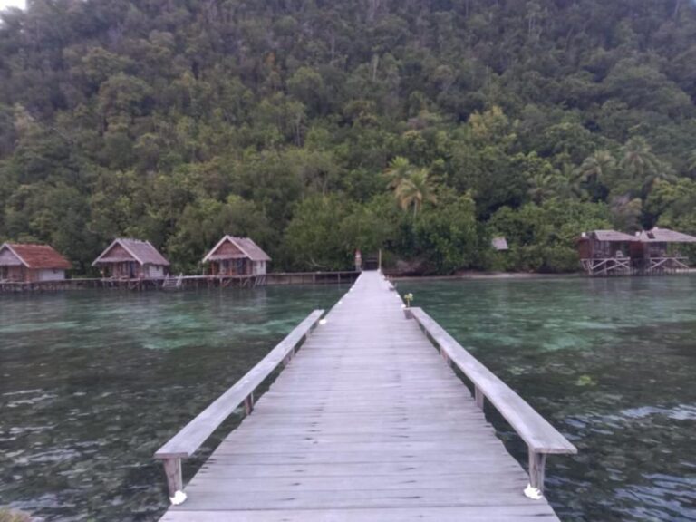 Ein langer Steg mit Bänken über ruhigem Wasser, gegenüber den Bungalows und dem bewaldeten Hügel