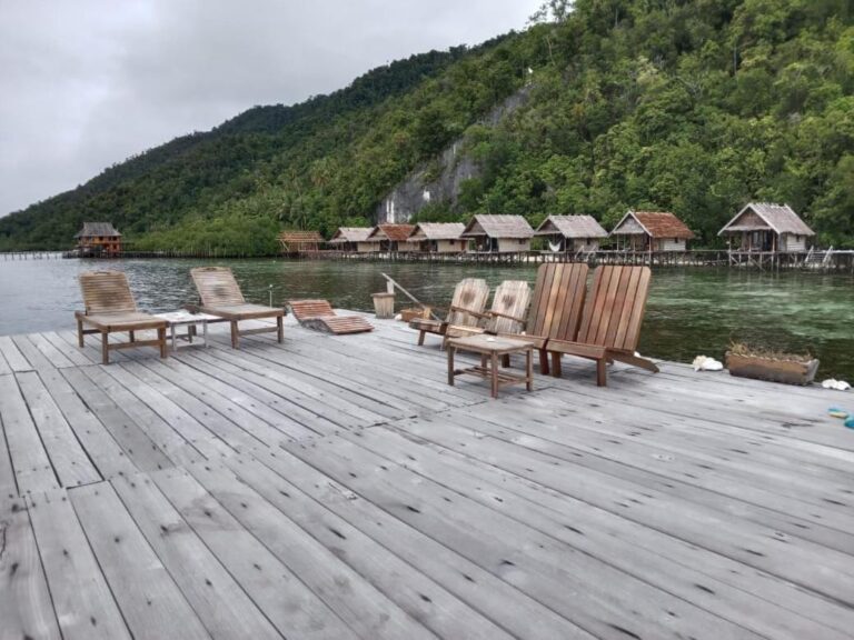 Liegestühle auf einem Steg bei grauem Wetter, vor den Bungalows und dem Hügel