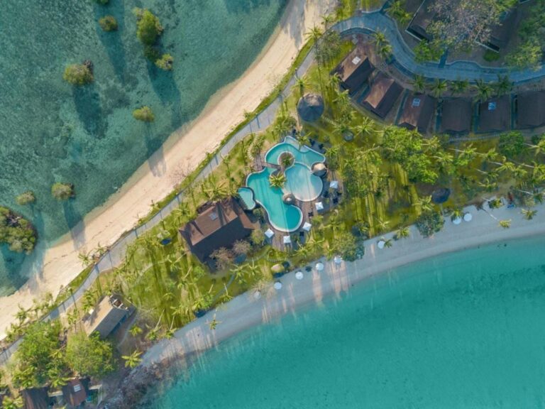 Blick von oben auf den Pool und die Dächer des Resorts, zwischen zwei Sandstränden