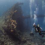 Der aufragende, mit Bewuchs bedeckte Bug der Giannis D, mit drei Tauchern am Fuß des Wracks in blauem Wasser voller Luftblasen
