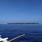Die flache, bewaldete Insel Balicasag am Horizont, mit ihrem weißen Leuchtturm und hellem Sandsaum, gesehen von einer Bangka, deren weiße Ausleger den Vordergrund einnehmen, unter wolkenlos blauem Himmel.