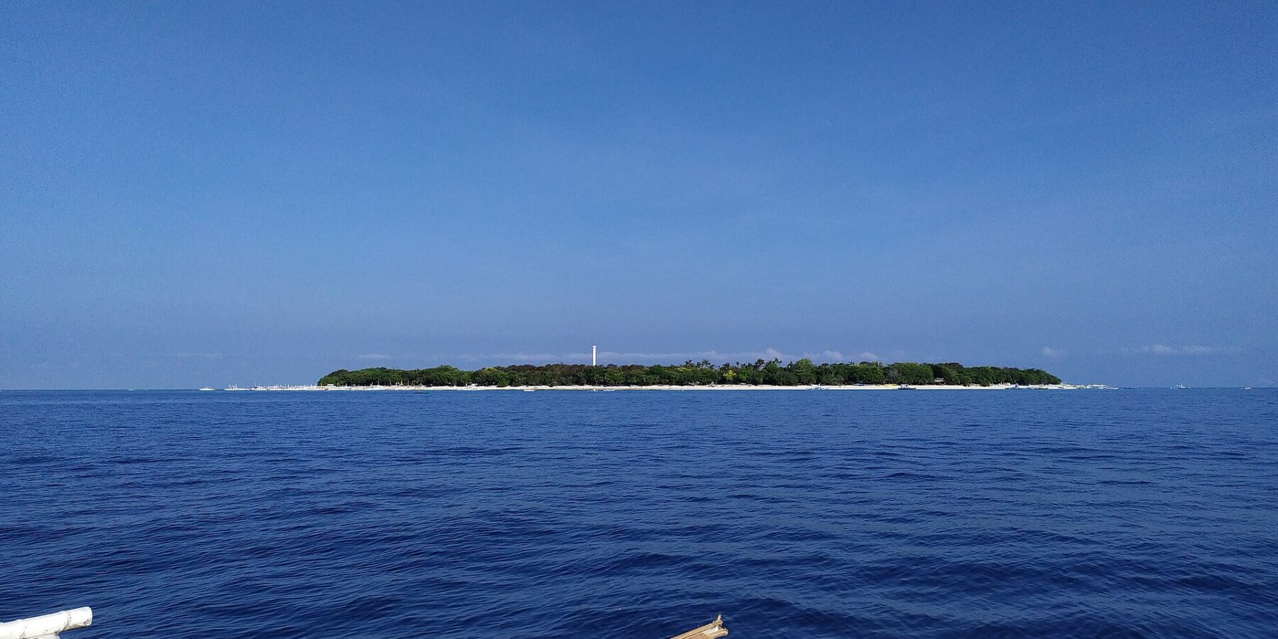 Die flache, bewaldete Insel Balicasag am Horizont, mit ihrem weißen Leuchtturm und hellem Sandsaum, gesehen von einer Bangka, deren weiße Ausleger den Vordergrund einnehmen, unter wolkenlos blauem Himmel.