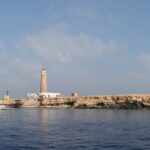 Big Brother vom Meer aus: eine flache, helle Insel mit ihrem Leuchtturm, davor ein weißes Safariboot vertäut