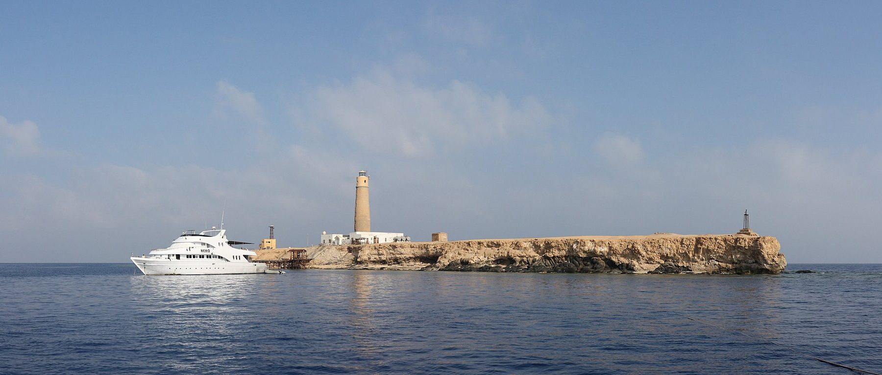 Big Brother vom Meer aus: eine flache, helle Insel mit ihrem Leuchtturm, davor ein weißes Safariboot vertäut