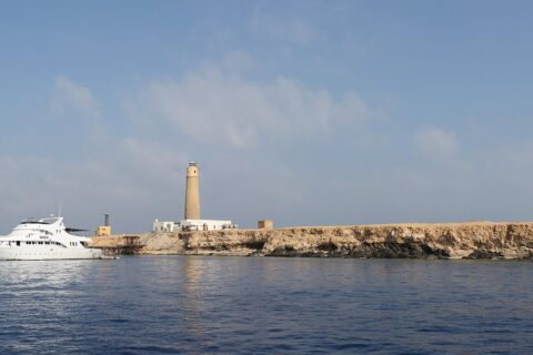Big Brother vom Meer aus: eine flache, helle Insel mit ihrem Leuchtturm, davor ein weißes Safariboot vertäut